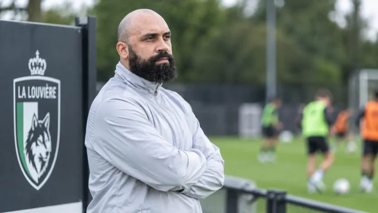 Anthony Vanden Borre kijkt langs een trainingsveld van La Louvière, met een clubembleem op een vast bord naast het veld.