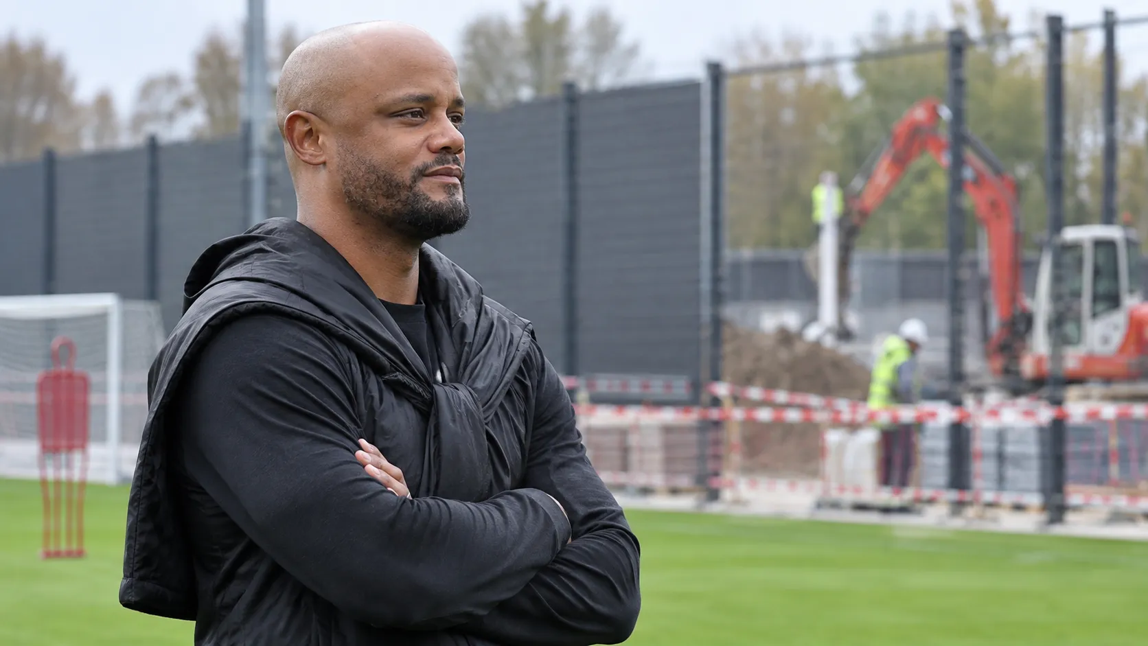 Vincent Kompany aan de rand van een trainingsveld bij een bouwzone met nieuwe afscherming.