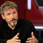 Een voetbalcommentator spreekt aan een studiodesk met microfoon.
