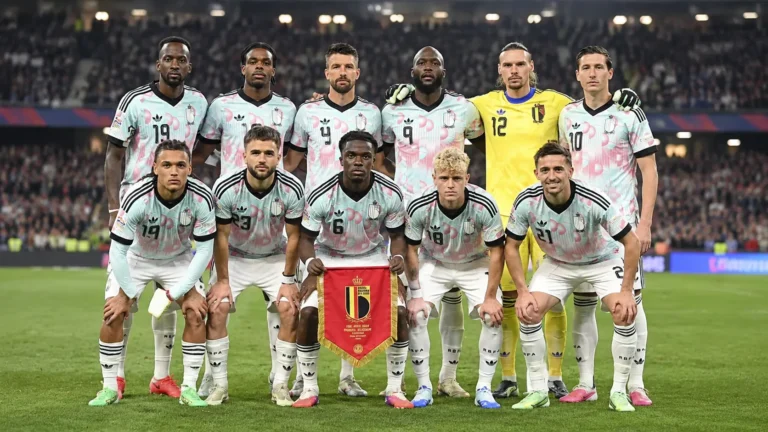Een groep Belgische nationale spelers poseert met een wimpel op het veld van een verlicht stadion.