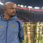 Een niet-spelende Bayern München-vertegenwoordiger staat naast een trofee aan de rand van het veld.