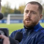 Gill Swerts spreekt ernstig langs een voetbalveld in een donkerblauwe sportjas.
