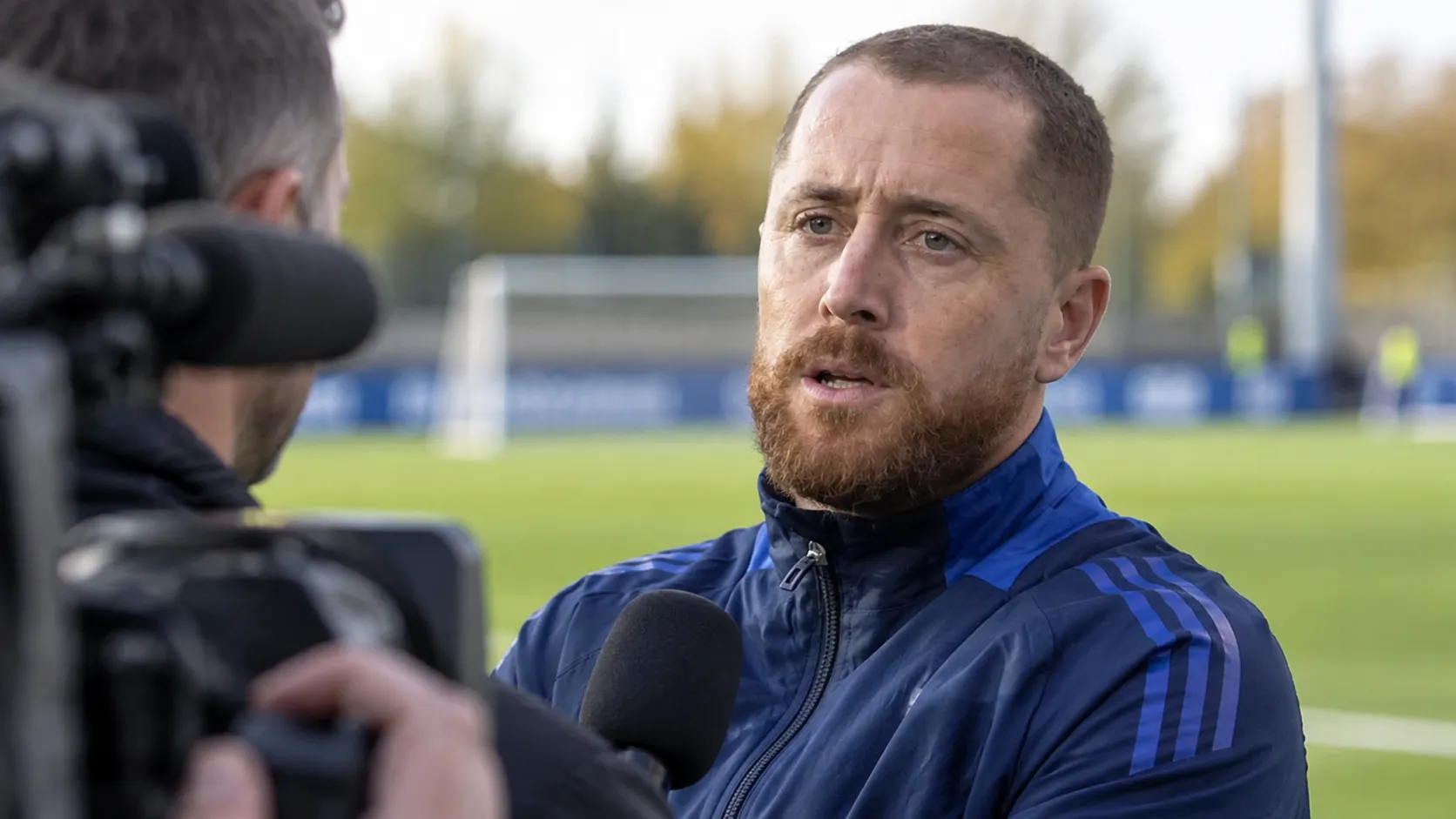 Gill Swerts spreekt ernstig langs een voetbalveld in een donkerblauwe sportjas.