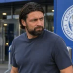 Russell Martin bij een stadioningang met een Leicester City-embleem op een vast bord achter hem.