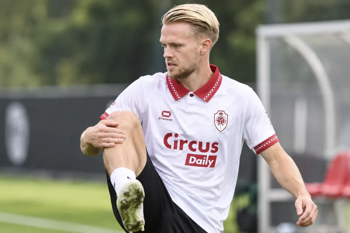 Een voetballer in een witte clubtrui warmt geconcentreerd op langs een trainingsveld.