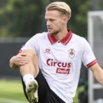 Een voetballer in een witte clubtrui warmt geconcentreerd op langs een trainingsveld.