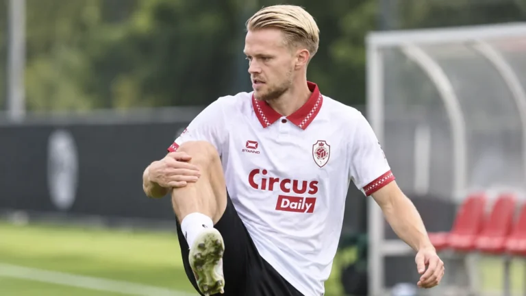 Een voetballer in een witte clubtrui warmt geconcentreerd op langs een trainingsveld.
