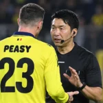 Een scheidsrechter spreekt op het veld met een speler in een geel tenue.