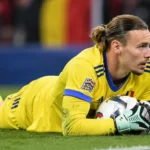 Maarten Vandevoordt in geel doelmanstenue met de bal na een lage redding op het veld.