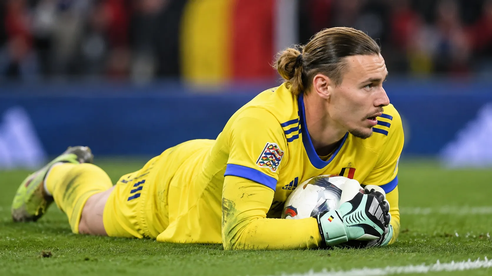 Maarten Vandevoordt in geel doelmanstenue met de bal na een lage redding op het veld.