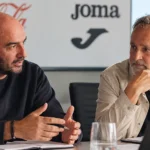 Twee niet-spelende clubvertegenwoordigers tijdens een overleg in een binnenruimte.