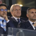 Niet-spelende clubvertegenwoordigers van Manchester City in de tribune van een stadion.