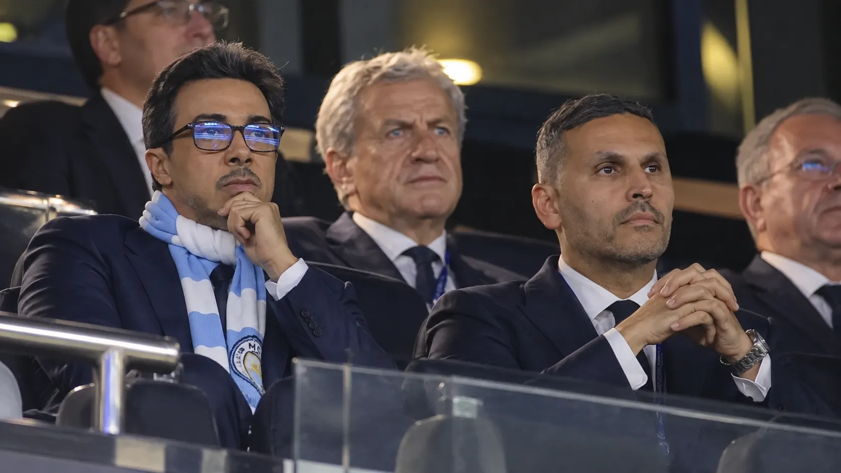 Niet-spelende clubvertegenwoordigers van Manchester City in de tribune van een stadion.