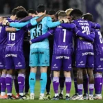 Een groep voetballers in paarse wedstrijdkleding vormt samen een hechte kring op het veld.