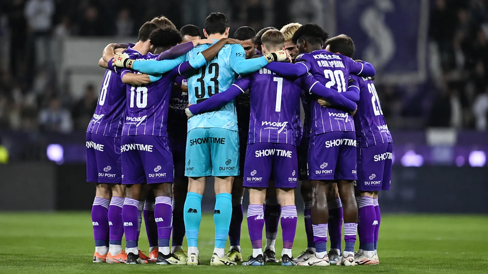 Een groep voetballers in paarse wedstrijdkleding vormt samen een hechte kring op het veld.