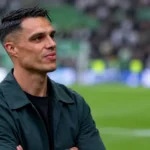 Joey Pelupessy in een donkere jas langs de rand van een voetbalveld.