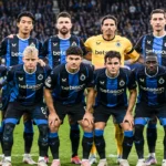 Een groep spelers van Club Brugge in blauw-zwarte tenues poseert in een stadion.