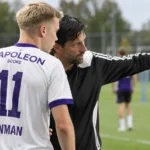 Een Anderlecht-trainer geeft langs het veld instructies aan een speler in een wit-paars shirt.