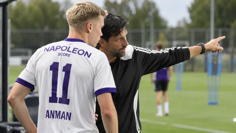 Een Anderlecht-trainer geeft langs het veld instructies aan een speler in een wit-paars shirt.