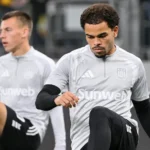 Twee voetballers in grijze opwarmkleding tijdens een moment naast het veld.