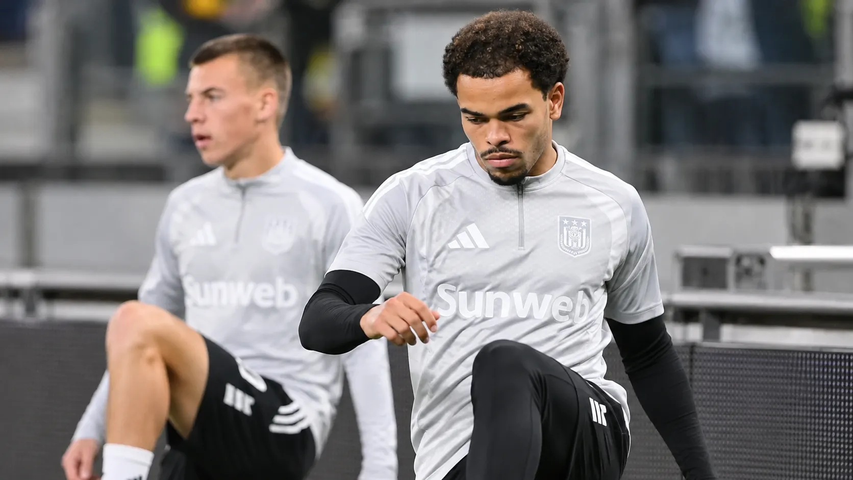 Twee voetballers in grijze opwarmkleding tijdens een moment naast het veld.