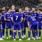 Een groep Anderlecht-spelers in blauw-paarse wedstrijdkleding verzamelt op het veld voor een wedstrijd.