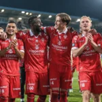 Een groep voetballers in rode tenues viert samen op het veld onder stadionlichten.