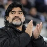 Een voormalige voetballer in een zwarte jas applaudisseert in een stadion.
