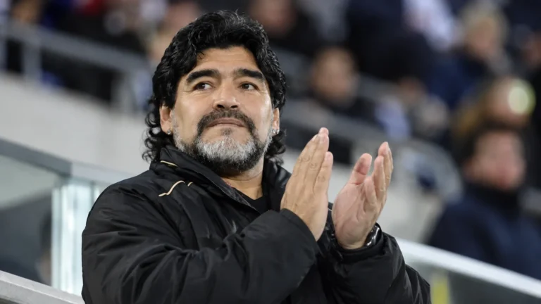 Een voormalige voetballer in een zwarte jas applaudisseert in een stadion.