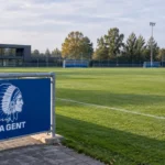 Leeg trainingsveld van KAA Gent met een blauw-wit clubembleem op een vast bord langs het veld.