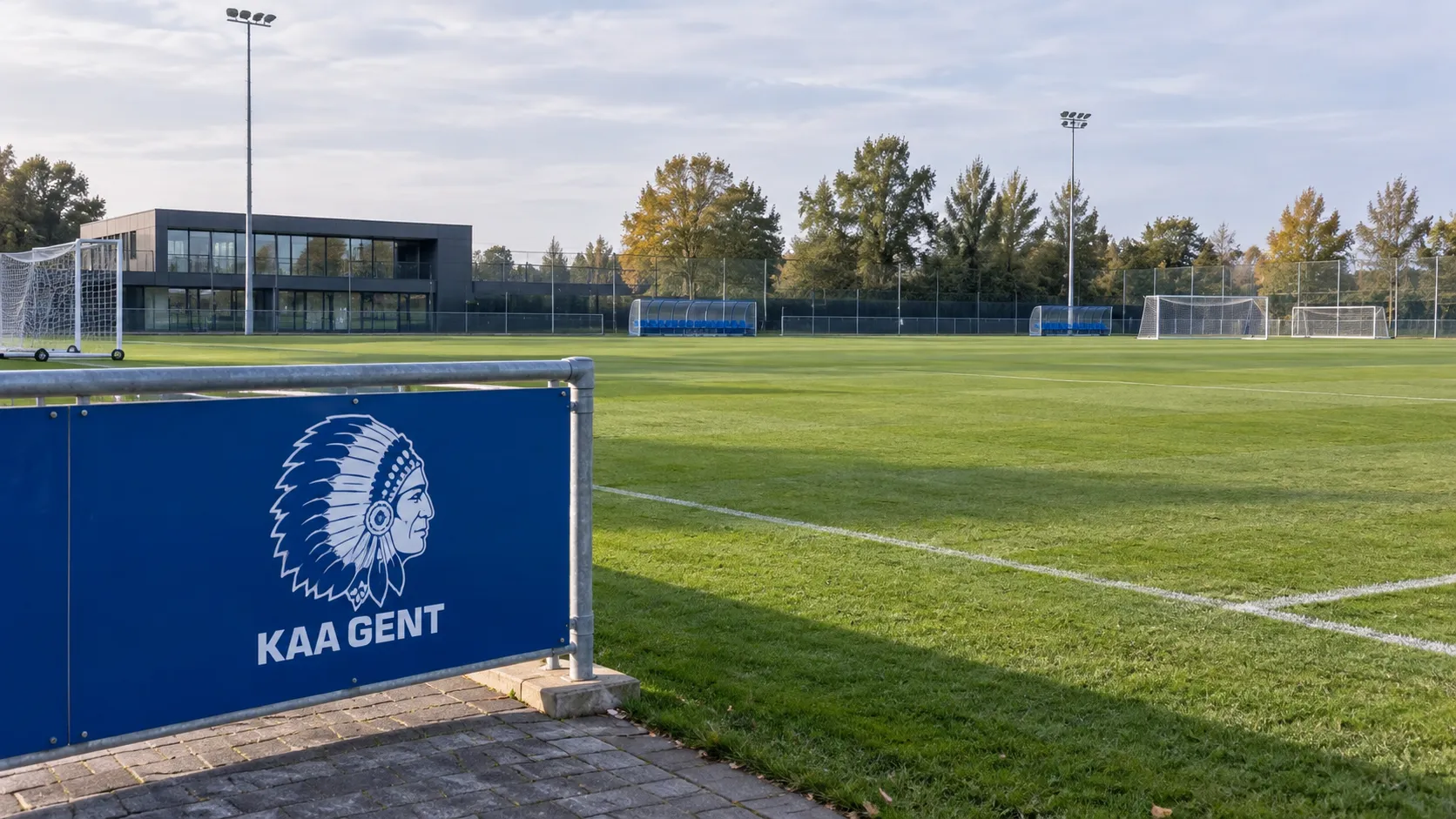 Leeg trainingsveld van KAA Gent met een blauw-wit clubembleem op een vast bord langs het veld.