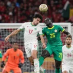 Meerdere voetballers in groene en witte tenues vechten een hoge bal uit voor doel.
