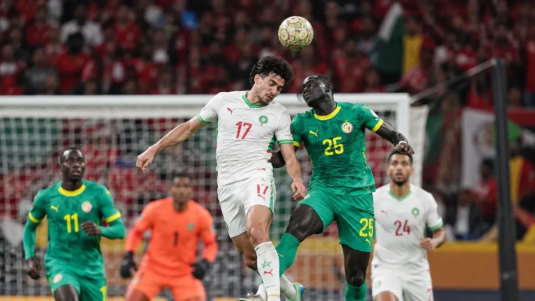 Meerdere voetballers in groene en witte tenues vechten een hoge bal uit voor doel.