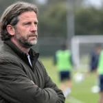 Een coach in een donkere jas volgt aandachtig de voorbereiding naast het voetbalveld.