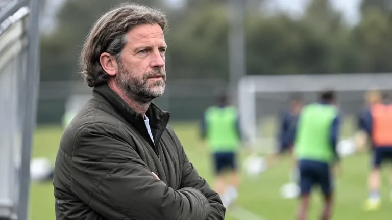 Een coach in een donkere jas volgt aandachtig de voorbereiding naast het voetbalveld.