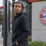 Michael Olise in neutrale sportkleding bij de ingang van het trainingscomplex van Bayern München.