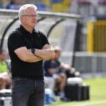 Een coach in een zwarte polo staat geconcentreerd in de technische zone naast het veld.