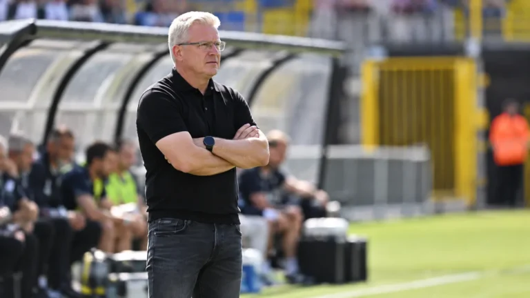 Een coach in een zwarte polo staat geconcentreerd in de technische zone naast het veld.