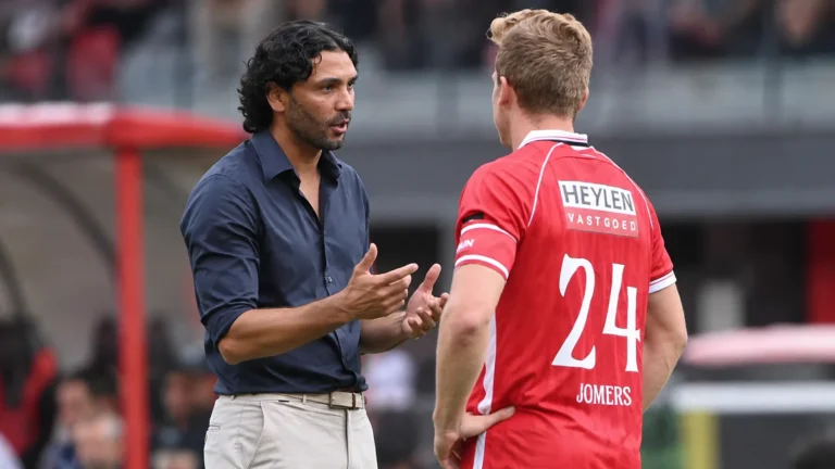 Een Antwerpse coach overlegt langs het veld met een speler in rood tenue.