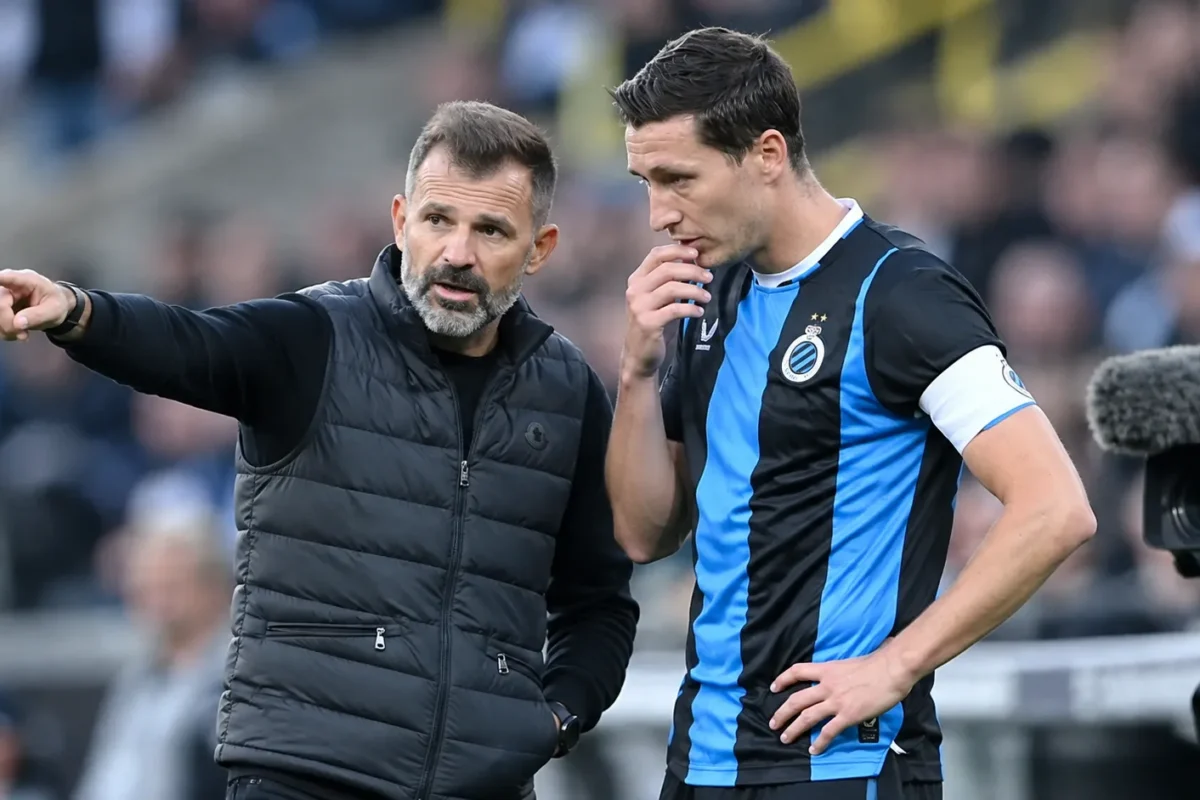 Een Club Brugge-coach spreekt langs het veld met een speler in blauw-zwart tenue.