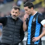 Een Club Brugge-coach spreekt langs het veld met een speler in blauw-zwart tenue.