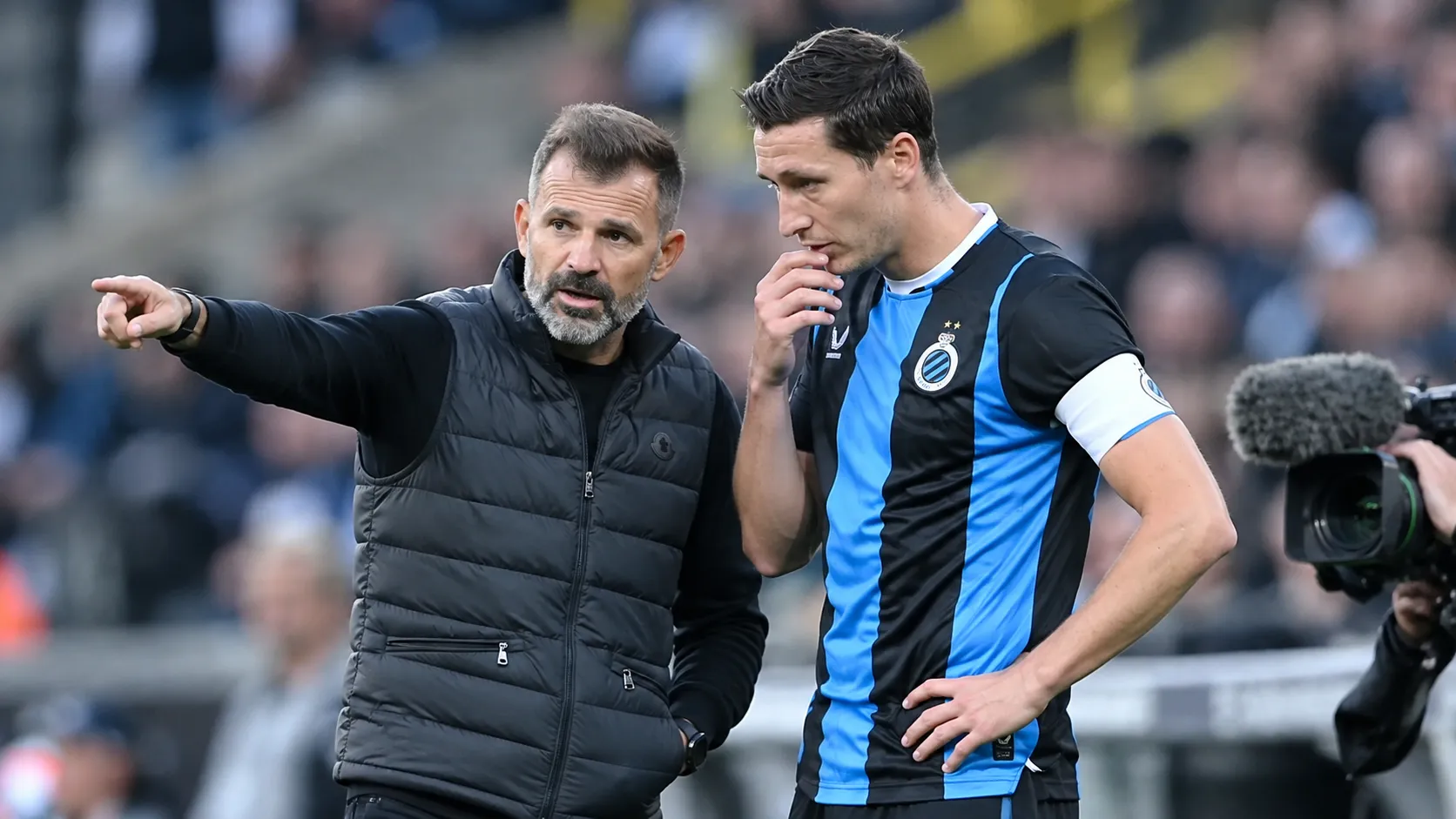 Een Club Brugge-coach spreekt langs het veld met een speler in blauw-zwart tenue.