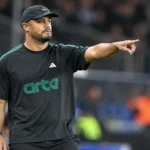 Een trainer in donkere sportkleding geeft aanwijzingen naast het voetbalveld.