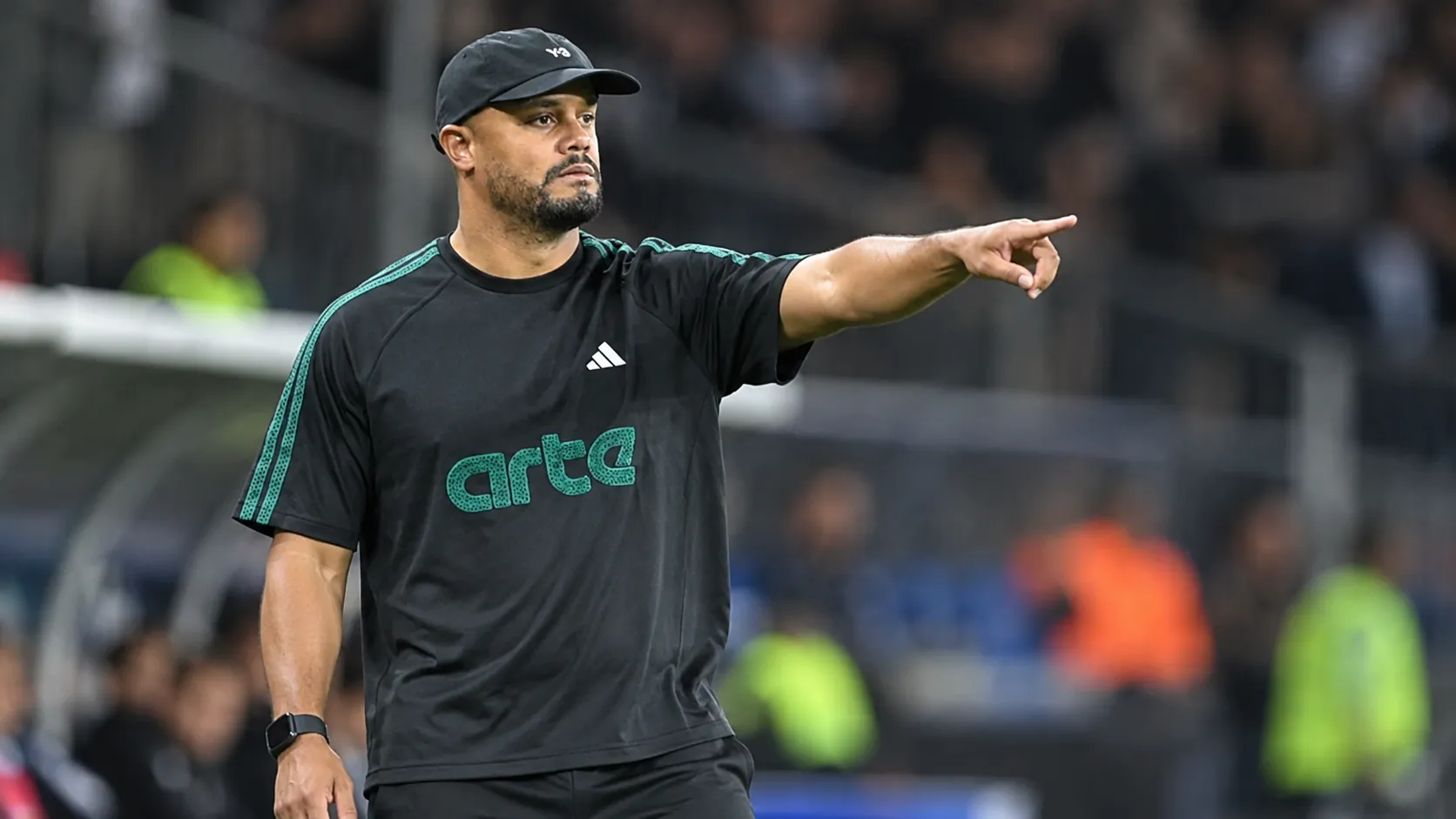 Een trainer in donkere sportkleding geeft aanwijzingen naast het voetbalveld.