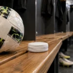Een voetbal en een gesloten, onbedrukt nicotinezakje op een houten bank in een lege kleedkamer.