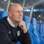 Een coach in een donkere jas zit in een blauwe stadiondug-out.