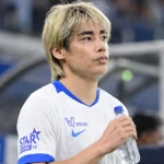Een speler van KRC Genk in een wit shirt met blauwe details tijdens een rustig moment naast het veld.