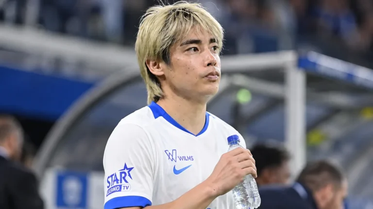 Een speler van KRC Genk in een wit shirt met blauwe details tijdens een rustig moment naast het veld.