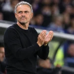 Een coach in donkere trui applaudisseert langs de zijlijn in een stadion.