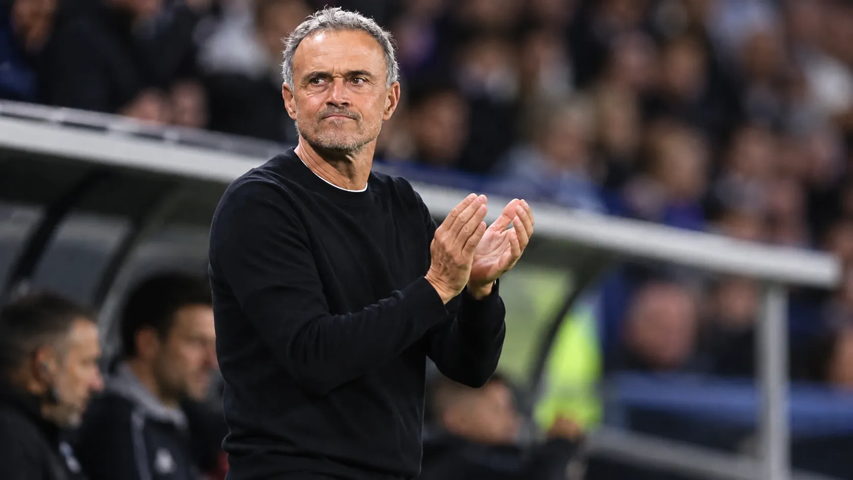 Een coach in donkere trui applaudisseert langs de zijlijn in een stadion.